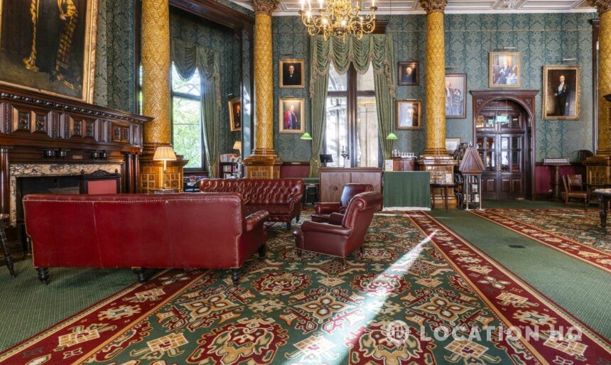 Luxurious historic lounge with rich decor, featuring tall golden columns, green damask wallpaper, and large windows draped in swag curtains