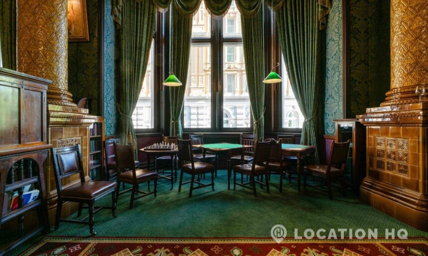 Luxurious historic lounge with rich decor, featuring tall golden columns, green damask wallpaper, and large windows draped in swag curtains